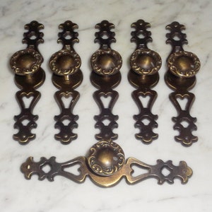 Może przedstawiać: Zestaw sześciu antycznych mosiężnych gałek do szuflad z ozdobnymi wzorami. Gałki mają wycięcie w kształcie serca i okrągły guzik z ozdobnym wzorem.