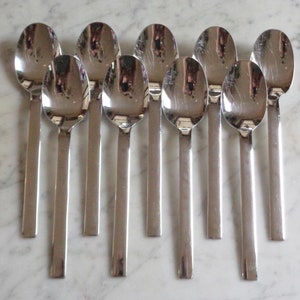 May include: Nine stainless steel spoons arranged on a marble surface. Each spoon has a rounded bowl and a long, rectangular handle. The spoons reflect the surrounding environment, creating a mirrored effect. The spoons are a set, perfect for dining.