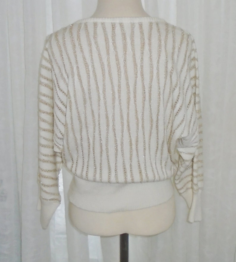 May include: A white sweater with a gold glitter stripe pattern. The sweater has a round neckline and long sleeves.