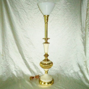 Könnte beinhalten: Eine hohe, goldene Stehlampe mit einem weißen, glockenförmigen Schirm. Die Lampe hat einen weißen und goldenen Sockel mit einem dekorativen Design.