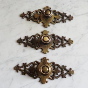 May include: Three ornate brass door knobs with a decorative, intricate design. Each knob has a round, raised center with a circular indentation.