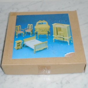 May include: A box containing a miniature dollhouse bedroom set. The set includes a bed, two chairs, a dresser, a nightstand, and a wardrobe. All the furniture is made of wood and painted a light yellow color.