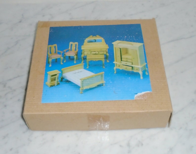 May include: A box containing a miniature dollhouse bedroom set. The set includes a bed, two chairs, a dresser, a nightstand, and a wardrobe. All the furniture is made of wood and painted a light yellow color.