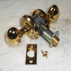 Op de afbeelding: Een gouden deurknop met een ronde knop en een vierkante sluitplaat. De knop heeft een sleutelgat en een kleine hendel aan de zijkant. De sluitplaat heeft het woord "Kwikset" erop gegraveerd.