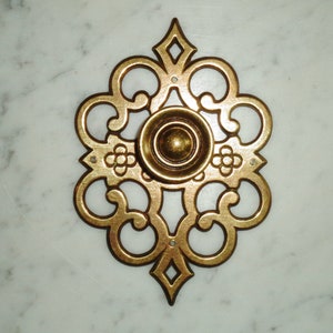 May include: A gold-toned metal cabinet knob with a decorative, ornate design. The knob has a round, raised center and a detailed, intricate pattern around the edges.