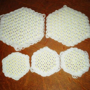 May include: A collection of white, hexagonal crochet items, including two larger trivets and three smaller coasters. Each piece features a floral pattern with yellow centers and a scalloped edge, all arranged on a wooden surface.