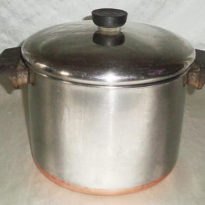 Puede incluir: Una olla de acero inoxidable con tapa y dos asas laterales. La olla tiene una base de color cobre y un pomo negro en la tapa. Diseñada para cocinar y preparar alimentos.