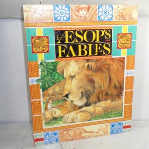 May include: A hardcover book titled "Aesop's Fables" with a colorful illustration of a lion and a mouse. The cover features a lion with a golden mane, lying on green grass. The title is in large, yellow letters.