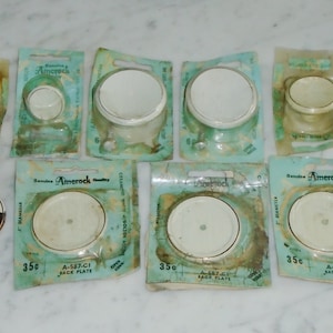 Puede incluir: Ocho placas de toma de corriente de cerámica blanca en envases individuales de plástico transparente. Cada envase tiene un fondo verde con el texto "Amerock" y "35c" impreso en él. El texto "A-537-C1 JACK PLATE" también está impreso en el envase.