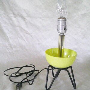 May include: A yellow ceramic bowl lamp with a black metal tripod base and a clear light bulb. The lamp has a black electrical cord with a plug.
