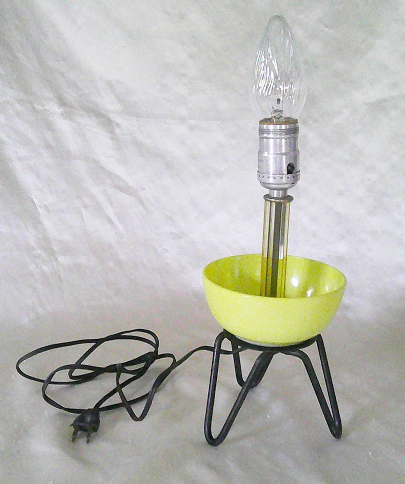 May include: A yellow ceramic bowl lamp with a black metal tripod base and a clear light bulb. The lamp has a black electrical cord with a plug.