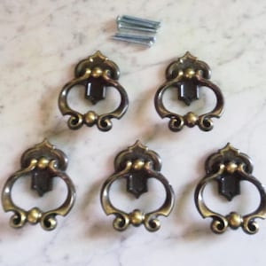 Puede incluir: Cinco tiradores de puerta de armario de latón antiguo con diseños ornamentados. Cada tirador tiene una abertura en forma de anillo y un patrón de volutas decorativo.