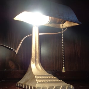 May include: A vintage gold-toned desk lamp with a shade that has a slight curve. The lamp has a pull chain and a decorative base.