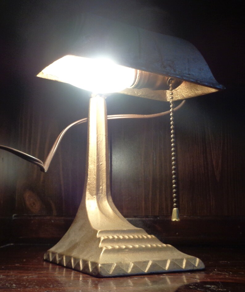 May include: A vintage gold-toned desk lamp with a shade that has a slight curve. The lamp has a pull chain and a decorative base.