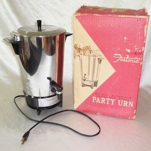 May include: A silver 30-cup automatic stainless steel party urn by Fostoria. The urn is on a white surface with its original box behind it. The box is red with white text that reads "30 CUP AUTOMATIC STAINLESS STEEL PARTY URN".