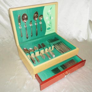 以下が含まれることがあります： 赤色の引き出しが付いたヴィンテージの木製カトラリーチェスト。銀色のカトラリーの完全なセットが含まれています。内側は緑色のフェルトで裏打ちされ、花の模様があります。チェストは開いており、スプーン、フォーク、ナイフが表示されています。