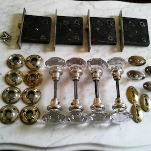 May include: A collection of antique door hardware, featuring four black metal mortise locks, various brass and silver-toned escutcheons, and five clear glass and metal door knobs. Screws and brass-toned rosettes are also present.