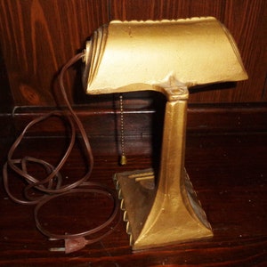 May include: A gold-colored metal desk lamp with a curved shade and a pull chain. The lamp has a cord and plug.