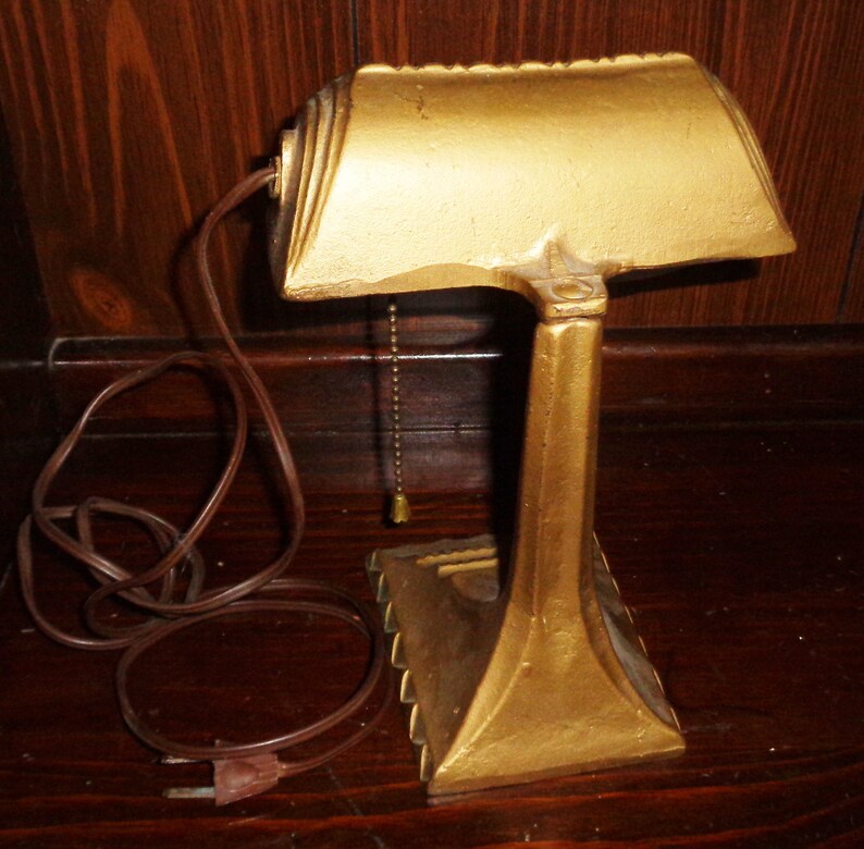 May include: A gold-colored metal desk lamp with a curved shade and a pull chain. The lamp has a cord and plug.