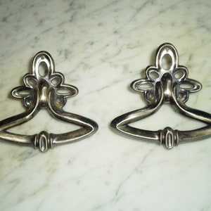 Könnte beinhalten: Zwei antike, silberfarbene Schubladengriffe mit verzierten Blumendesigns. Die Griffe sind tropfenförmig mit abgerundeter Oberseite und gebogenem Boden.