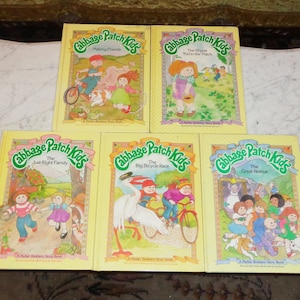 Op de afbeelding: Vijf vintage Cabbage Patch Kids-verhalenboeken met gele covers. Titels zijn onder meer "Making Friends", "The Shyest Kid in the Patch", "The Just Right Family", "The Big Bicycle Race" en "The Great Rescue". Elk boek bevat kleurrijke illustraties.