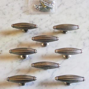 May include: Eight silver-toned cabinet knobs with a ribbed design. The knobs are oval-shaped with a flat base and a small, round handle on top.