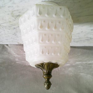 May include: A vintage white glass light fixture with a tiered, geometric design. The fixture has a decorative brass base and a frosted appearance. The light fixture is designed to be mounted on a ceiling.