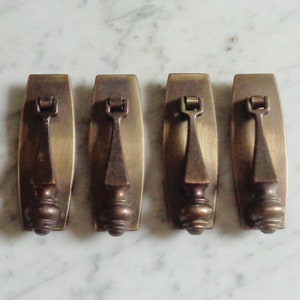 以下が含まれることがあります： 長方形で尖った装飾的なハンドルが付いた、アンティークな真鍮製のキャビネットプルが4つ。プルは、わずかに古びた、くすんだ仕上げになっています。