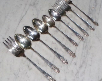 May include: A set of eight silver-plated spoons and forks with ornate handles. The utensils are arranged in a row on a white marble surface.