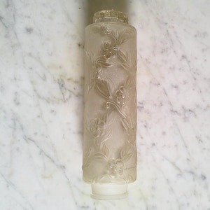 May include: A cylindrical, frosted glass lampshade with an embossed floral design. The shade is clear with a textured surface and a slightly flared base. The top has a decorative, ridged cap.