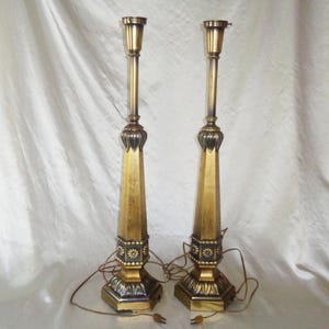 May include: A pair of gold-toned table lamps with a geometric design. The lamps have a tall, slender base and a decorative finial at the top.