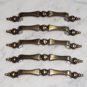 Könnte beinhalten: Fünf antike Messing-Schrankgriffe mit dekorativem Design. Die Griffe haben eine abgerundete Form mit einer leichten Biegung und einen dekorativen Knopf an jedem Ende.