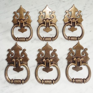 Puede incluir: Seis tiradores de armario de latón antiguo con un diseño decorativo ornamentado. Cada tirador tiene una manija de anillo y un detalle de cuerda trenzada.