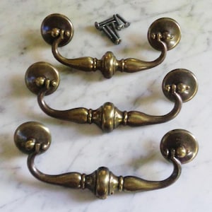 May include: Three antique brass drawer pulls with matching screws. Each pull has a curved handle and round backplates. The brass has a warm, aged patina, suggesting a vintage style. Suitable for furniture restoration or adding a classic touch.