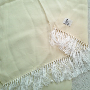 May include: A pale yellow fabric with white fringe trim. The fringe is attached with small white beads. A tag in the upper right corner reads "Tops" with a small hat logo.