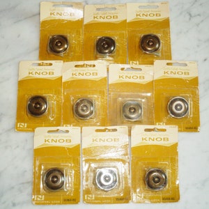 Op de afbeelding: Tien verpakkingen messing kastknoppen, elk met een transparant plastic venster dat de knop laat zien. De verpakkingen zijn geel met zwarte tekst die "Quality Cabinet Hardware Knob" en "National Lock" luidt.