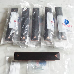 Peut inclure: Six emballages individuels de poignées de tiroir en bronze foncé avec un motif triangulaire et un seul emballage d'une poignée de tiroir en bronze foncé plus longue avec un motif triangulaire. Les emballages sont étiquetés "Hickory Hardware".