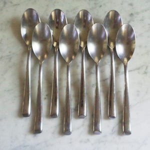 Puede incluir: Una colección de ocho cucharas de acero inoxidable dispuestas sobre una superficie de mármol. Cada cuchara tiene un mango largo y delgado y un cuenco redondeado. Las cucharas son reflectantes, mostrando el entorno.