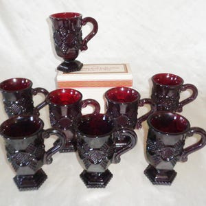 Op de afbeelding: Set van acht robijnrode glazen bekers met een reliëfpatroon. De bekers hebben een voet en een handvat. De bekers zijn in een cirkel op een wit oppervlak gerangschikt. De bekers maken deel uit van de Cambridge Glass Collection.