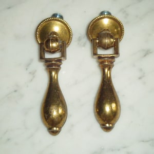 Könnte beinhalten: Zwei antike Schubladengriffe aus Messing. Jeder Griff hat eine kreisförmige Rückplatte mit einem seilartigen Rand, ein rechteckiges Verbindungsstück und einen tropfenförmigen Griff. Auf einer weißen Marmoroberfläche.