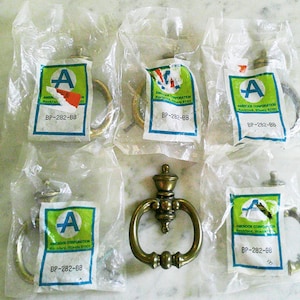 May include: Five brass ring pulls in clear plastic packaging and one loose brass ring pull. Each package features a white label with a green square, a blue letter A, and the text Amerock Corporation, Rockford, Illinois, and BP-282-BB.