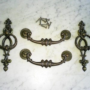 May include: Antique bronze-toned drawer pulls and decorative pulls with screws. The drawer pulls have a curved handle design with ornate detailing. The decorative pulls have a ring design with a fleur-de-lis top. Hardware for furniture restoration.