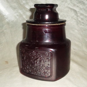 Op de afbeelding: Een donkerrode pot met een deksel, in antieke stijl. De pot heeft een rechthoekige vorm met een hart en bloemenmotief in reliëf op de voorkant. De deksel is plat en zit bovenop de pot. De pot is van glas.