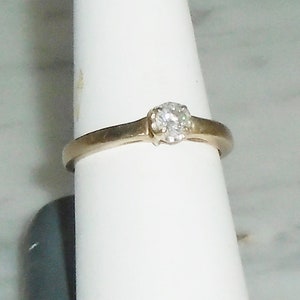 May include: A gold-toned ring with a single, round, clear diamond set in a prong setting. The ring is displayed on a white surface, showcasing the simple elegance of the jewelry.