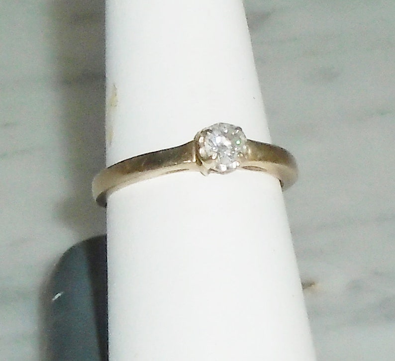 May include: A gold-toned ring with a single, round, clear diamond set in a prong setting. The ring is displayed on a white surface, showcasing the simple elegance of the jewelry.