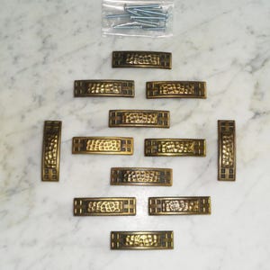 Op de afbeelding: Tien antieke messing ladegrepen met een gehamerde afwerking en een geometrisch ontwerp. De grepen zijn symmetrisch gerangschikt op een wit marmeren oppervlak.