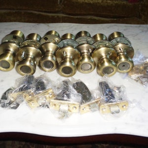 May include: A set of ten gold-toned door knobs with a keyhole and a small, clear plastic window. The knobs are arranged in two rows on a white marble surface. There are also several black plastic pieces in the image.