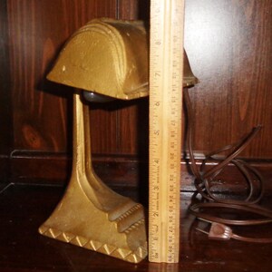 May include: A gold-colored metal desk lamp with a geometric design. The lamp has a round shade and a long, slender base. The lamp is plugged in and has a brown cord.