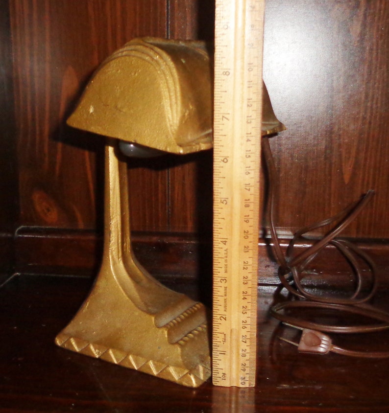 May include: A gold-colored metal desk lamp with a geometric design. The lamp has a round shade and a long, slender base. The lamp is plugged in and has a brown cord.
