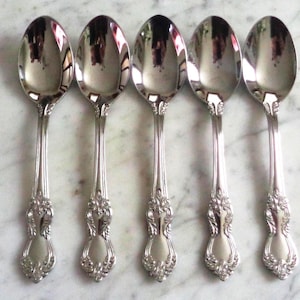 Può includere: Cinque cucchiai d'argento con dettagli floreali ornati sui manici sono disposti in fila. I cucchiai hanno una superficie riflettente e lo sfondo è una superficie chiara, simile al marmo. I cucchiai sono probabilmente per mangiare.